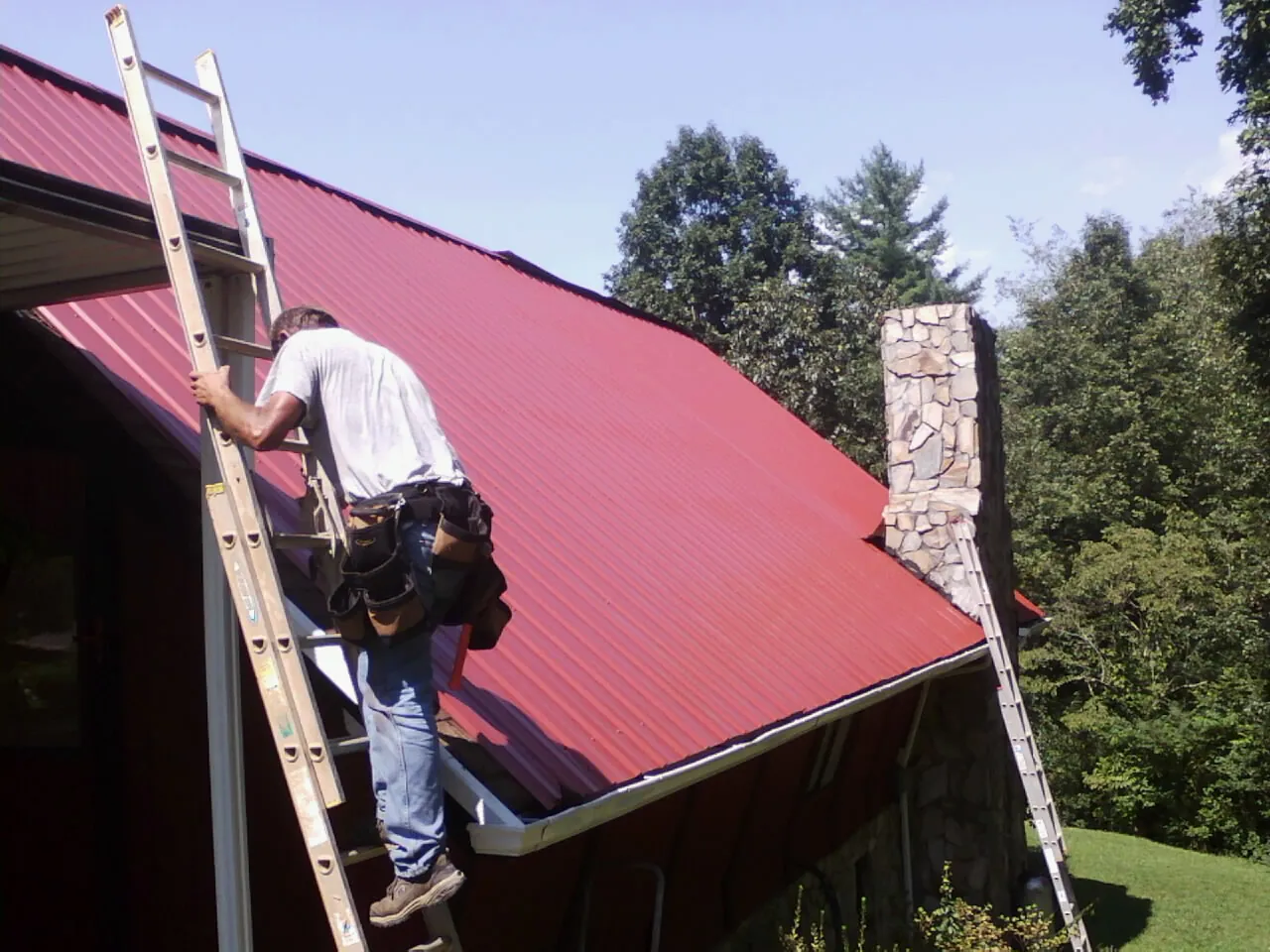 Roofer on a ladder beside a red metal roof and stone chimney — Soffit & Fascia Repair by Roofing Brothers in Kirkersville